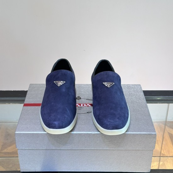 Front toe-up view of Prada Men's blue suede slip-on loafers showing enamel triangle logo in blue and white