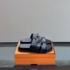 Hermes Jackson Leather Sandals In Black - Paired View in black and silver Pair of Hermes Jackson Leather Sandals In Black resting on an orange designer box in black and silver